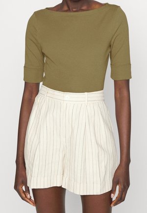 Femme portant un haut vert olive à manches coudes associé à un short crème taille haute avec des rayures verticales fines et une ceinture boutonnée.