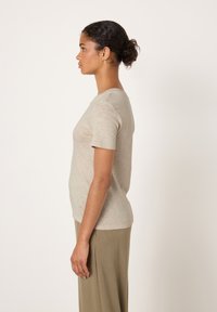 Young woman with curly hair in a bun wearing a beige short-sleeve shirt and olive-green pants, standing in profile against a white background.