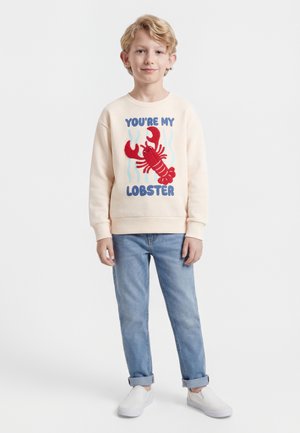 Young boy wearing cream sweatshirt with red lobster graphic and text "YOU'RE MY LOBSTER," blue jeans, and white slip-on shoes, standing.