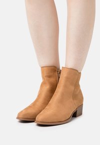 Beżowe botki zamszowe z gładkim wykończeniem, zaokrąglonym noskiem i niskim, piętrowym obcasem. Zamek błyskawiczny z boku, brak widocznych wzorów.