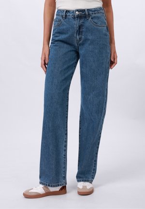 Woman wearing high-waisted blue straight-leg jeans with front pockets and white sneakers, standing on a plain background.