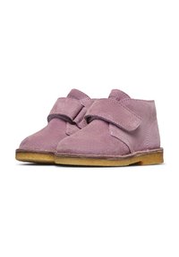 Un paio di scarpe per bambini in camoscio rosa con ampie cinghie in velcro e suole testurizzate color marrone chiaro, presentate su uno sfondo bianco.