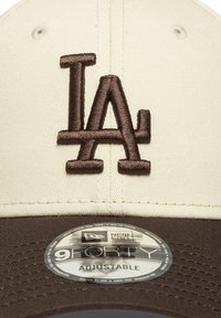 Gorra de béisbol ajustable con la parte delantera en crema y el visera marrón, con un logo bordado en marrón que dice "LA" y una etiqueta redonda que dice "9FORTY."