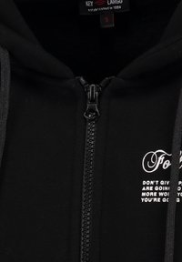 Sudadera negra con cremallera fabricada en un material suave. Presenta una cremallera en la parte delantera, capucha con cordón y detalles en texto blanco en el lado izquierdo. Etiqueta de tamaño visible.