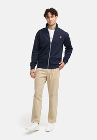 Navy zip-up jakke laget av myk stoff med krage; har en rød logo på venstre bryst. Kombinert med beige bukser og hvite sneakers.