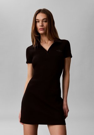 Young woman with long brown hair wearing a short-sleeve black polo dress, standing against a plain light gray background.