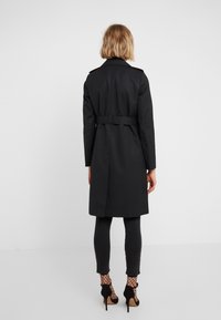 Žena s krátkými blond vlasy má na sobě páskový černý trench coat, černé kalhoty, punčochy z rybí kůže a černé vysoké podpatky, stojí na bílém pozadí.