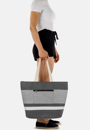 Graue und weiße gestreifte Stofftasche mit einem Reißverschlusstasche, flachem Boden und dicken Canvas-Trägern, gehalten von einer Person in lässiger Kleidung.