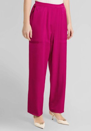 Donna che indossa pantaloni fucsia a vita alta e gambe larghe, abbinati a décolleté bianchi con punta appuntita e dettaglio a catena dorata.