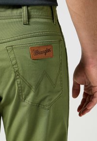 Groene Wrangler-broek met een leren logo-label op de achterzak, voorzien van een gestructureerde stof en zichtbare stiksels.