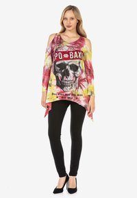 Farbiger Cold-Shoulder-Top mit einem Totenkopf-Graphic, ausgestattet mit einem Batikmuster in Rot und Gelb, kombiniert mit schwarzen Slim-Fit-Hosen und High Heels.