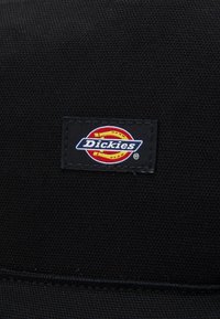 Étiquette en tissu noire avec un logo rouge, jaune et bleu comportant le texte "Dickies" et un design graphique, cousue sur un matériau texturé.