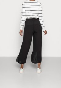 Zwarte wijde broek met zijsplits; gecombineerd met een witte gestreepte blouse met lange mouwen. De outfit wordt compleet gemaakt met witte sneakers.