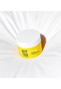 Pot cylindrique jaune étiqueté "NIP+FAB BARRIER CERAMIDE FIX CLEANSING BALM" avec un couvercle blanc, posé sur un fond blanc texturé.