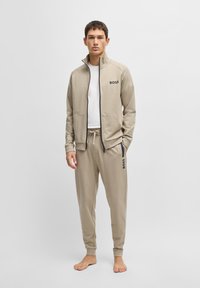 Beige trainingspak met een ritsjack en broek met een trekkoord, beide met zwarte "BOSS" logo-accenten. Glad materiaal, getailleerd ontwerp, casual.