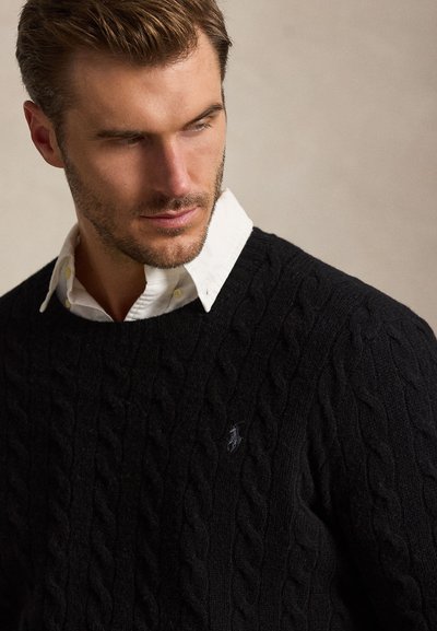 Giovane uomo con capelli corti e castani guarda verso il basso, indossando una camicia bianca con colletto sotto un maglione nero lavorato a trecce.