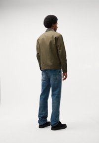Olivno-zeleni plašč z bež ovratnikom, oblečen preko modrih denim hlač. Plašč ima sproščen kroj in minimalističen dizajn. Vidi se črne čevlje.