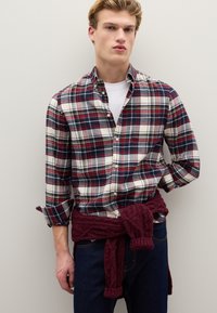 Camisa de cuadros en azul marino, rojo y crema con botones, mangas remangadas; lleva un suéter de punto burdeos anudado alrededor de la cintura. Jeans oscuros y camiseta blanca interior.