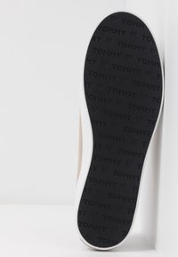 Sohle eines beigen Sneakers mit weißer Zwischensohle, schwarzer Außensohle mit geprägt wiederholtem "TOMMY"-Schriftzug und herzförmigen Mustern für besseren Halt.