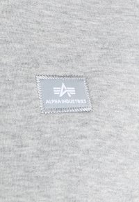Tissu gris à texture lisse avec un patch cousu portant l'inscription « ALPHA INDUSTRIES », surmonté d'un dessin de trois lignes horizontales.