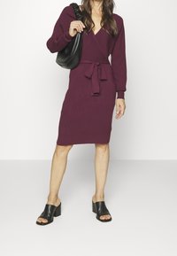 Robe en maille bordeaux foncé avec un design portefeuille, manches longues et ceinture à nouer à la taille. Associée à des mules noires à talon bloc et bout ouvert.