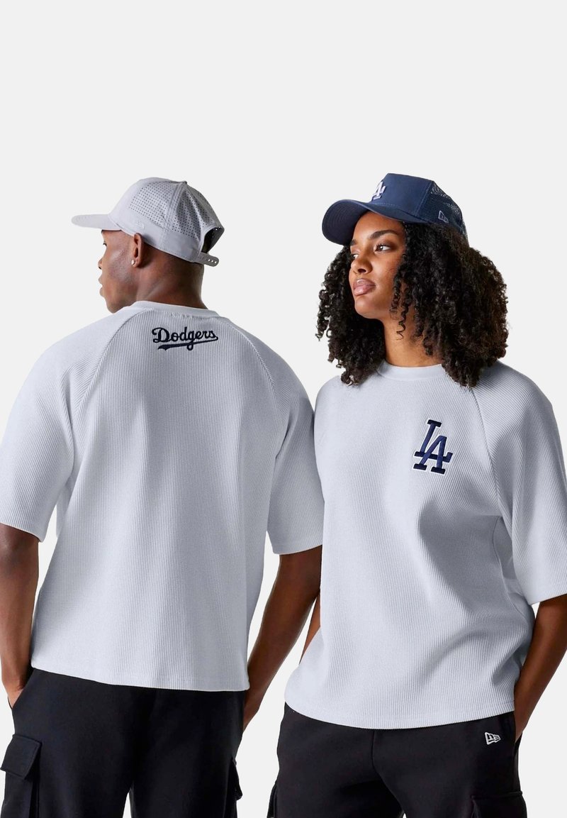 Chemise blanche à manches courtes côtelée avec "Dodgers" brodé en bleu marine au dos. Deux personnes portant des casquettes, l'une bleu marine et l'autre en filet gris.