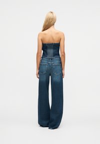 Femme aux longs cheveux blonds porte un haut en denim sans bretelles et un jean bleu à jambes larges, debout de dos sur un fond clair.