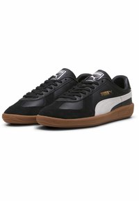 Pár černých tenisek Puma s bílým bočním pruhem, gumovou podešví, černými tkaničkami a zlatým logem Puma na boku a na jazyku.