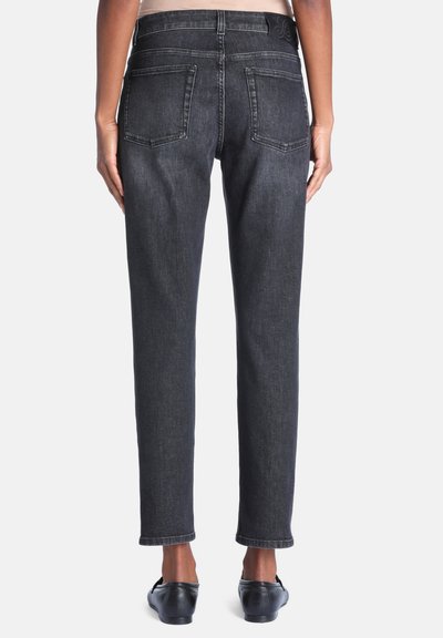 Vue arrière d'une personne portant un jean gris foncé droit et des chaussures noires sans lacets sur fond uni.