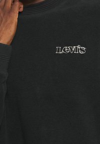 Persona che indossa un felpa nera Levi's, mano che tocca parzialmente il volto, logo Levi's ricamato in bianco sul petto.