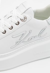 Sneaker platform bianca realizzata in materiale sintetico liscio. Presenta il logo "Karl" in rilievo, accenti in argento e design con lacci e occhielli rotondi.