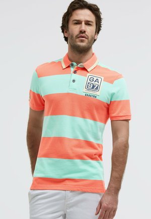 Mann trägt ein kurzärmeliges Poloshirt mit breiten horizontalen Korallen- und Minzstreifen, steht vor einem schlichten hellen Hintergrund.