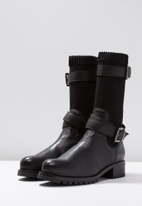 Las botas de cuero negro cuentan con un eje de tela acanalada, correas con hebillas planas y un tacón grueso con una suela texturizada que proporciona agarre.