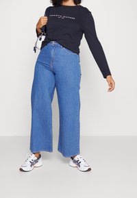 Jeans larghi blu con vita alta, abbinati a una maglietta a maniche lunghe nera con un logo. Sneakers bianche con accenti multicolori.