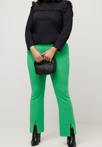 Femme portant un pantalon vert vif avec des fentes sur le devant, un haut noir à manches longues avec un col volanté, des bottines noires, tenant un petit sac à main noir.