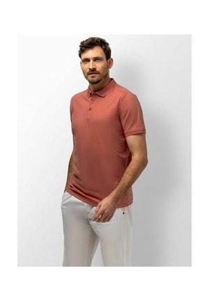 Man with short brown hair and beard wearing a rust-colored polo shirt and white pants standing against a light gray background.