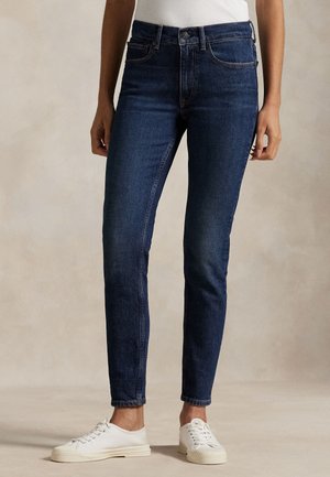 Person trägt dunkelblaue Slim-Fit-Jeans, weiße Sneakers und ein weißes Oberteil und steht vor einem einfarbigen hellen Hintergrund.