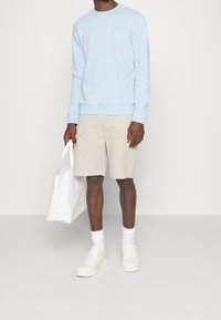 Ljusblå sweatshirt med ribbade ärmar, beige shorts, vita strumpor och ljusa sneakers. En vit axelremsväska hålls i ena handen.