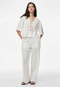 Femme portant une tenue en tissu à œillets blancs avec un haut en V noué, un pantalon à jambes larges et des sandales ouvertes noires, se tenant les mains dans les poches.
