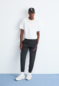 Mand iført sort baseball cap, hvid T-shirt, sorte joggers med rød lynlåsdetalje, hvide sokker og hvide sneakers står op ad en hvid væg.