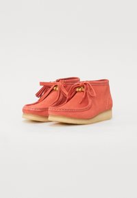 Botas de tobillo de ante de color coral con acentos de borlas, puntera redonda y suela de goma beige. Presentan costuras decorativas y detalles de nudos con cuentas.