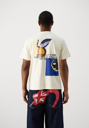 Cream t-shirt with graphic prints in blue and yellow, short sleeves, relaxed fit, paired with dark denim pants featuring red accents.