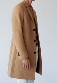 Abrigo de lana de camello con corte ajustado, que presenta un cuello con muescas, seis botones negros y un acabado liso y texturizado.