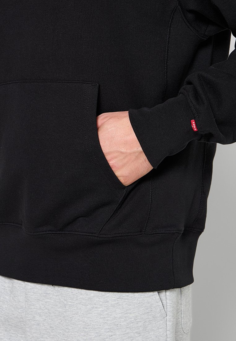 Schwarzer Baumwollpullover mit Kängurutasche und gerippten Bündchen. Ein kleines rotes Etikett ist am Ärmel sichtbar. Weicher Stoff, lässiges Design.
