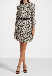 Vit klänning med svart blommönster, volangärmar, lagerdesign och ett svart bälte med cirkulär spänne. Sandalerna är svarta med ankelremmar.