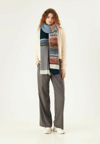 Mehrfarbiger gestrickter Schal mit blauen, grauen und burgunderroten Streifen. Weiche Textur und unterschiedliche Breiten, getragen über einem cremefarbenen Pullover.