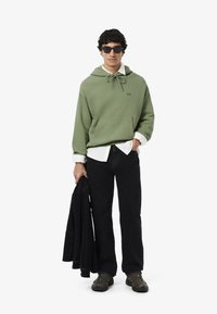 Felpa verde oliva con cappuccio e cordoncino, tasca frontale; indossata sopra una camicia bianca. Jeans neri e giacca nera. Scarpe da trekking marroni. Occhiali da sole.
