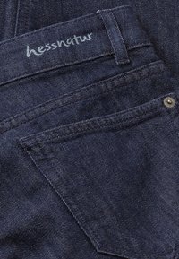 Pantalones de mezclilla oscura con una etiqueta hessnatur cosida en blanco. La tela tiene una textura suave con un sutil brillo y acentos de metal.