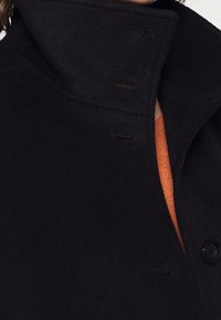 Zwarte woljas met een hoge kraag, knoopdetails en een getextureerde stof. Een oranje top is zichtbaar eronder.