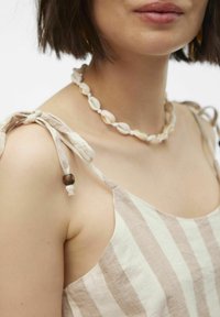 Top met bandjes, gemaakt van lichte stof met lichtbruine en crèmekleurige strepen, gekenmerkt door geknoopte schouderbanden en een schelpketting als accent.